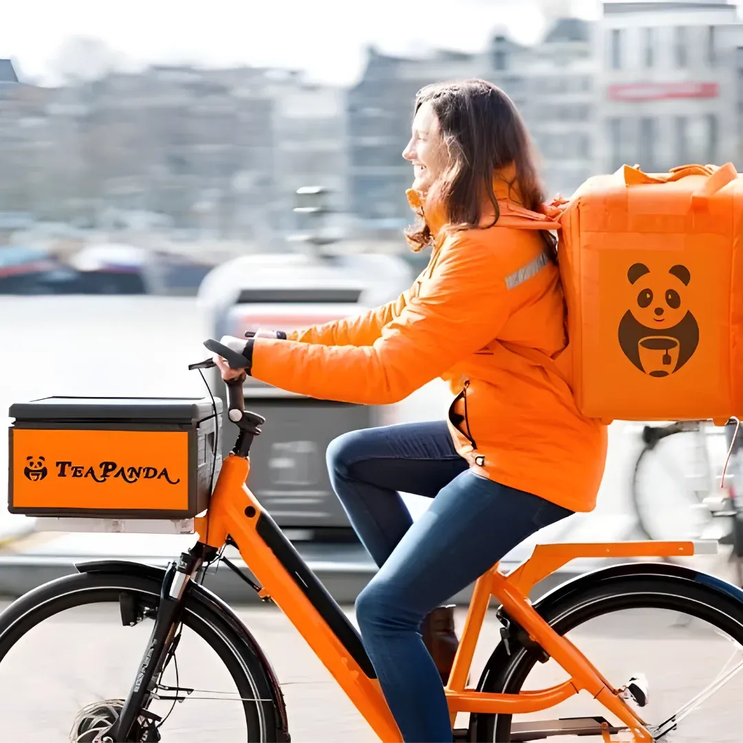 A woman riding a bicycle with a box on the back of it. She is wearing an orange jacket and blue pants, and the background is slightly blurred.