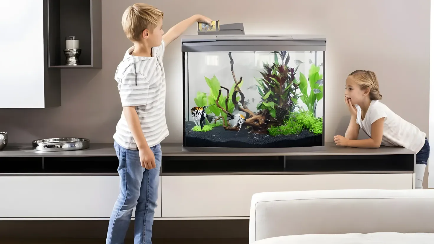 A boy and a girl looking at a fish tank in a living room. The aquarium is filled with water and plants, and there is a sofa in the bottom right corner of the room. On the left side of the image, there are bowls and other objects on the table, and in the background there are cupboards and a wall.