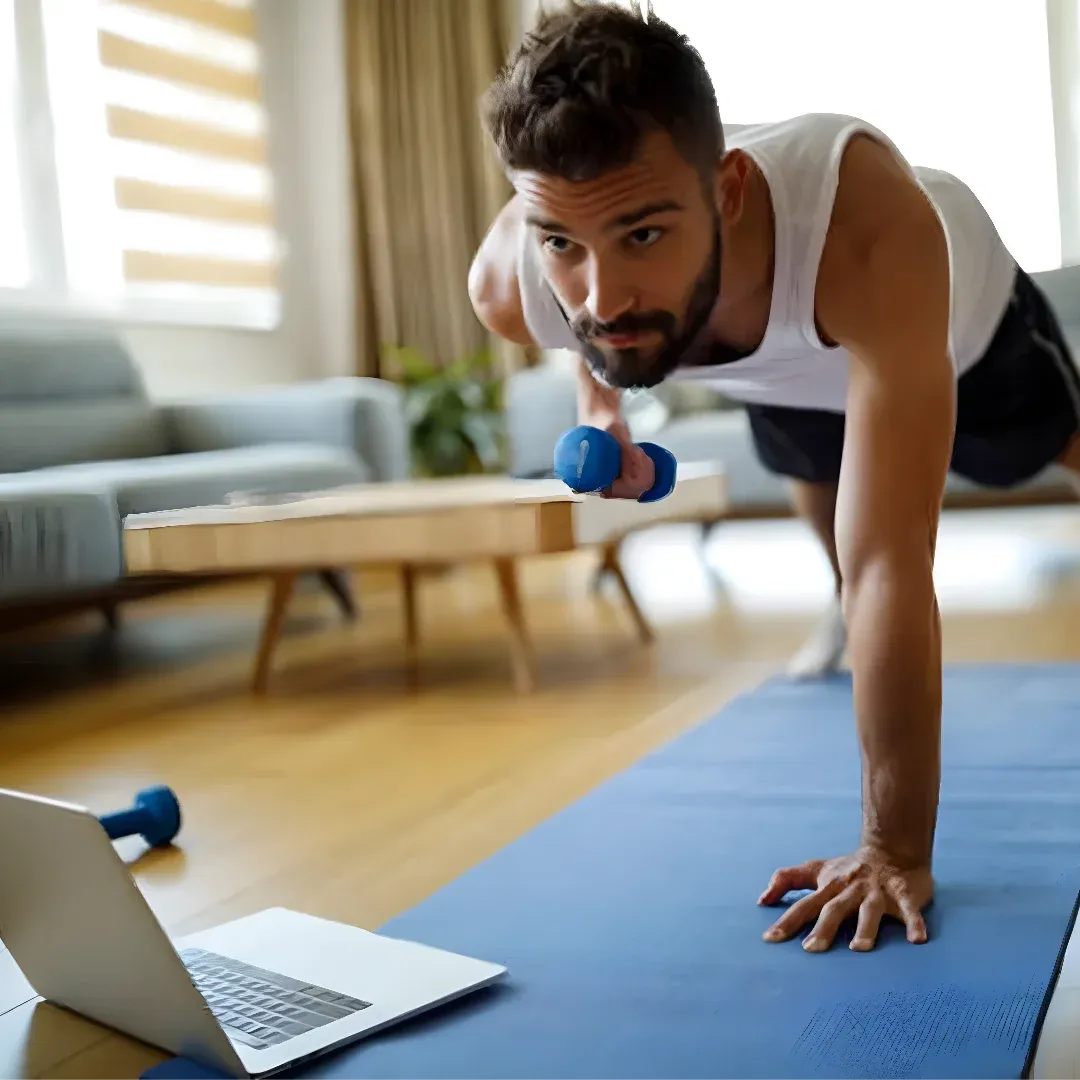 A man doing push ups on a yoga mat with a laptop in front of him. He is wearing a white t-shirt and black shorts, and is holding a pair of dumbbells in his hands. In the background, there is a sofa, a table, a plant, and windows with curtains.