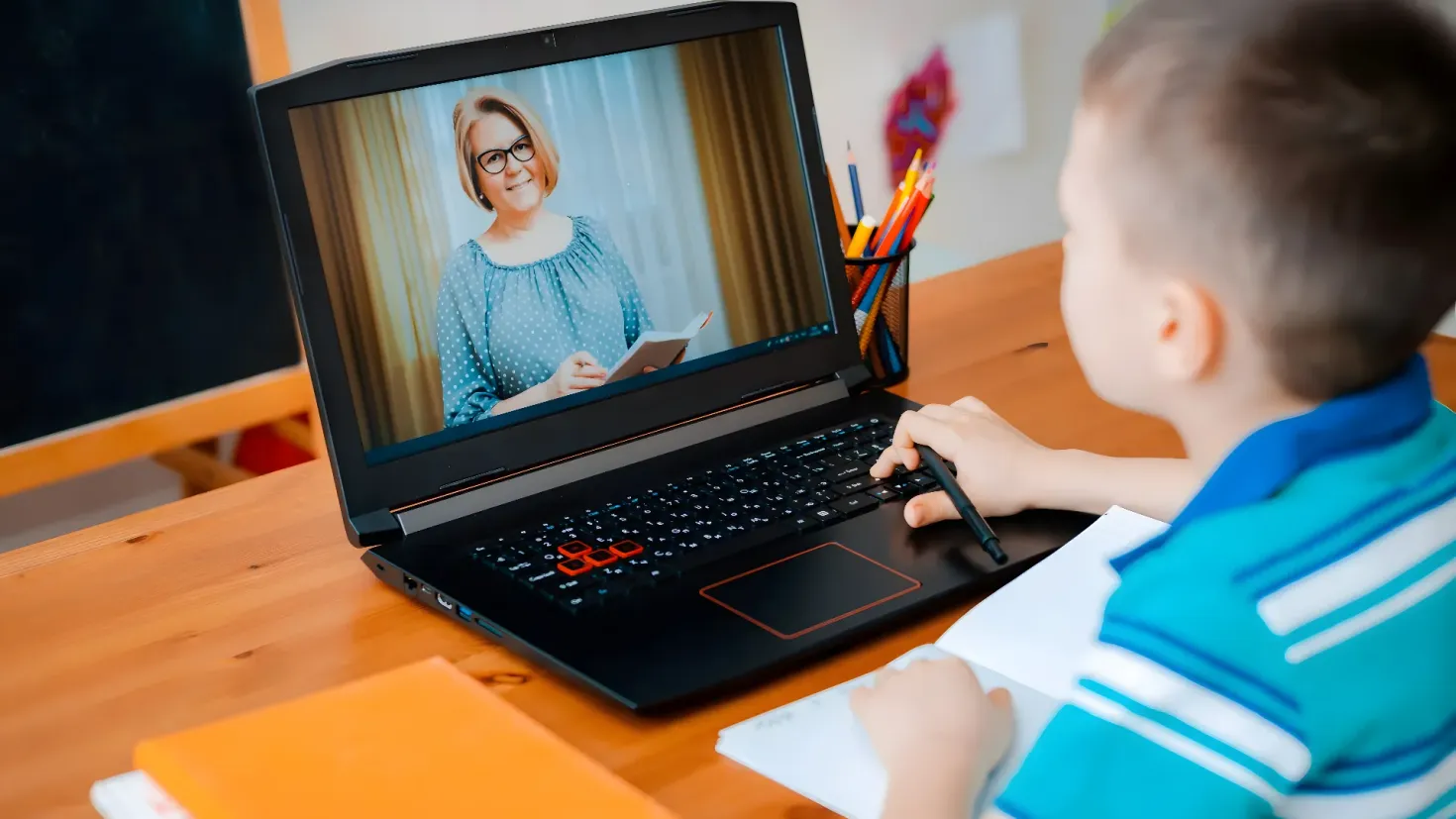A young boy sitting at a desk with a laptop computer in front of him. He is holding a pen in his hand and there are books and a pen stand with pencils on the desk. On the laptop screen, there is a picture of a woman and some text. In the background, there are frames attached to the wall.