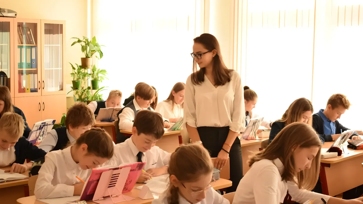 A group of children sitting at desks in a classroom. There are books, pens, and other objects on the desks, and a woman standing in the center of the room. On the left side of the image, there is a cupboard filled with books, and in the background, there are houseplants and a wall.