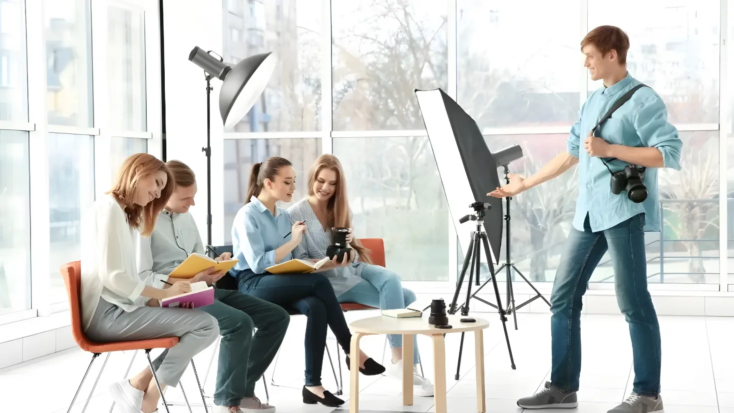 A group of people sitting around a table in front of a camera, with a man standing and holding a camera in his hand. On the table there are books and other objects, and in the background there are lights, stands, and glass doors. Through the glass doors, we can see trees, buildings, and the sky, suggesting that the group is in a photography studio.