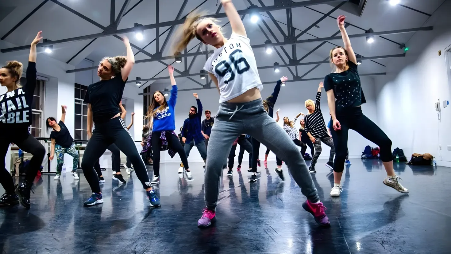 A group of people dancing in a dance studio. We can see a woman in the center of the group, surrounded by other people dancing on the floor. On the right side of the image, there is a wall with a door, and on the left side, there are windows and a door. At the top of the roof, we can see metal rods and lights, and there are bags and other objects scattered around the room.