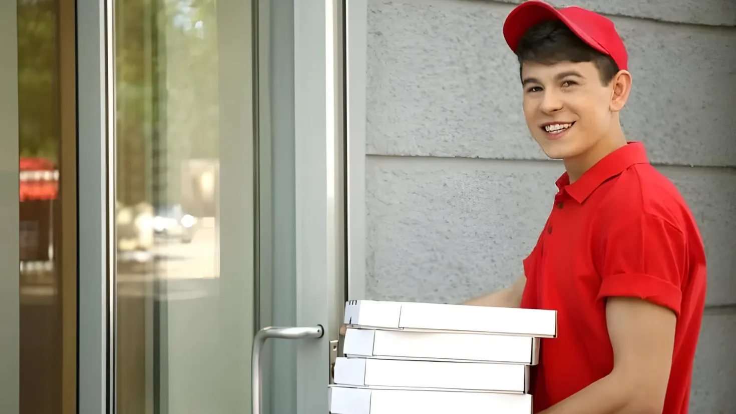 A young man wearing a red t-shirt and a red cap, holding a stack of pizza boxes in front of a glass door with a handle. In the background, there is a wall.