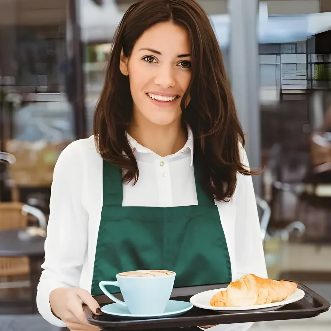 A waitress standing and smiling while holding a tray with a cup of coffee and a croissant on it. The background is slightly blurred, suggesting that the focus is on the waitress and her breakfast.