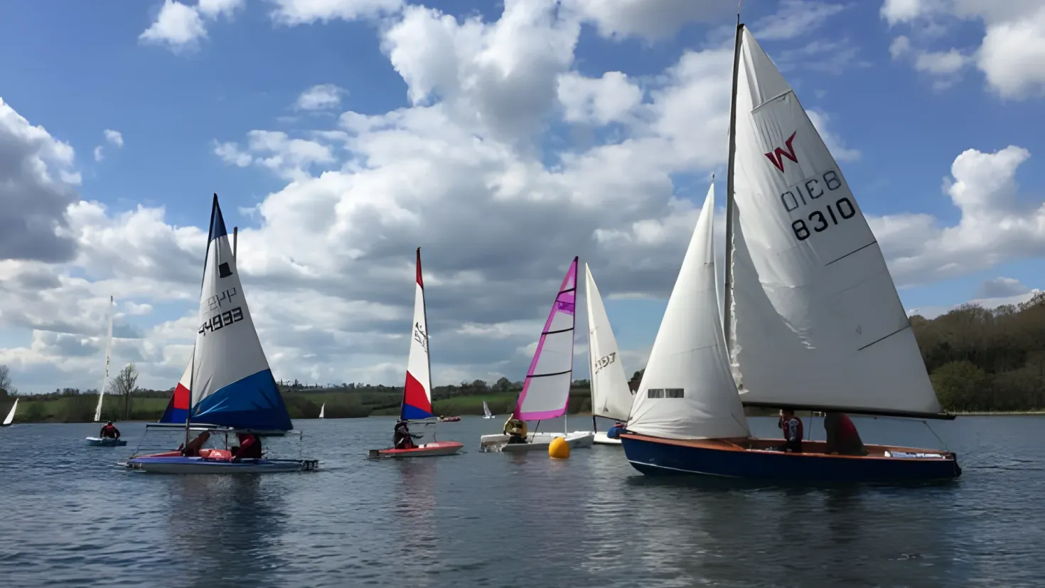 A group of sailboats floating on top of a body of water, with people in the boats. In the background, there are trees and a sky with clouds.