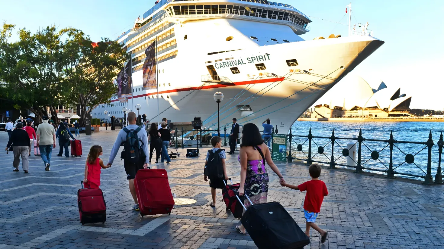 A group of people walking down a sidewalk next to a cruise ship, with a railing on the right side of the image. In the background, there is a body of water, trees, light poles, buildings, and a clear blue sky. Some of the people are carrying bags, suggesting they are on a journey.