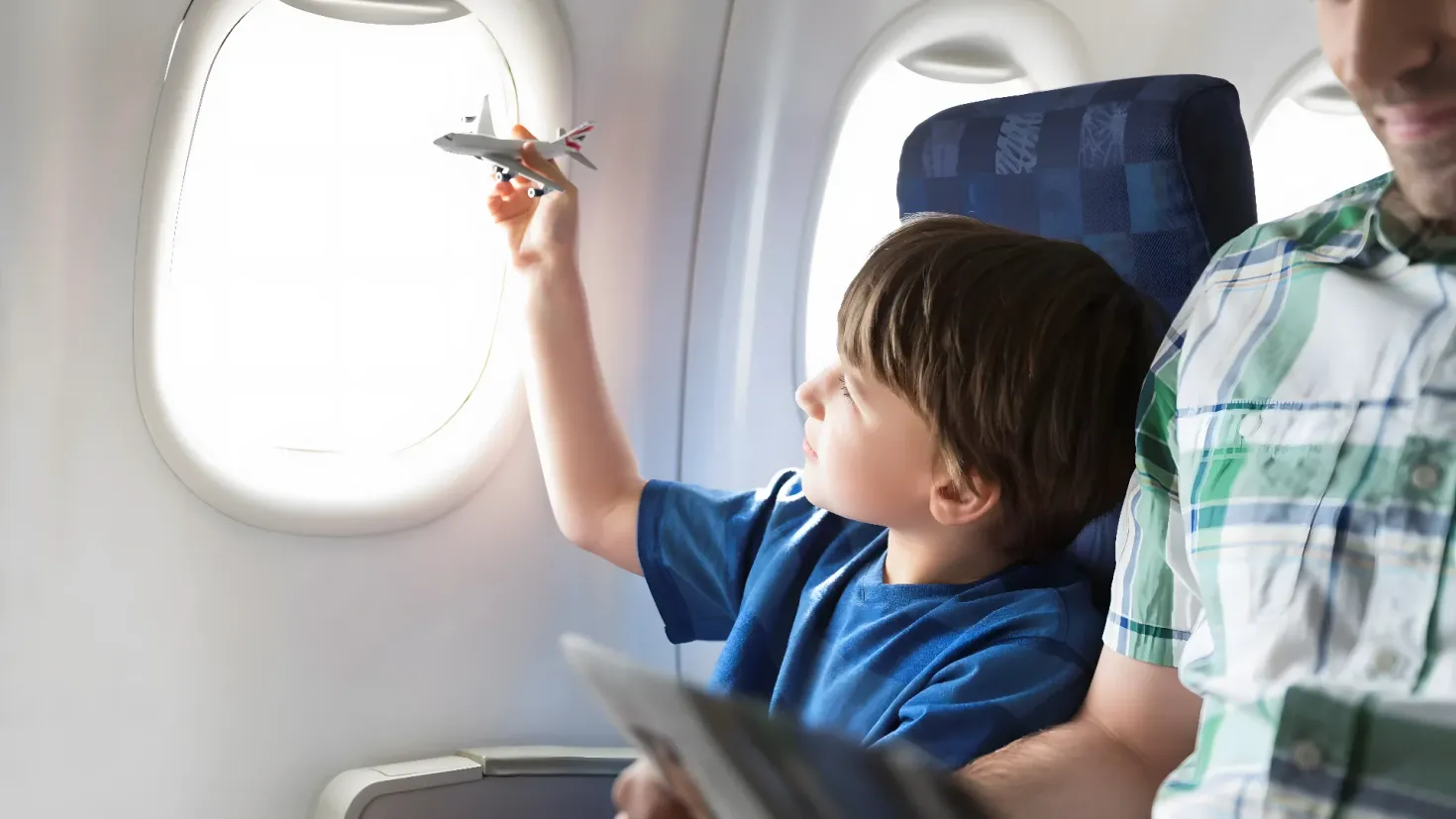 A father and son sitting in an airplane, with the father holding a toy airplane in his hand and the son looking out the window.
