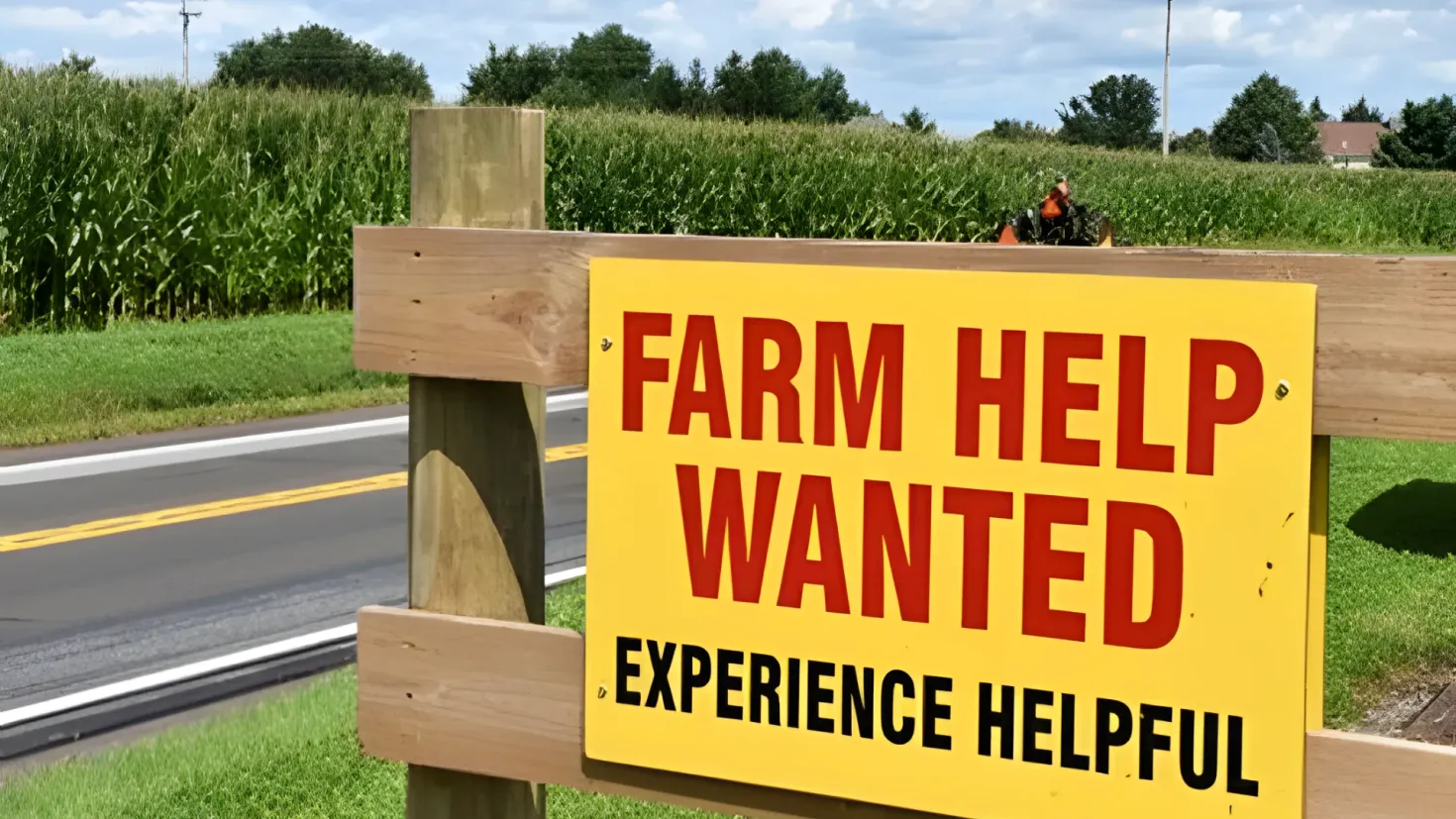 A person riding a motorbike on the road surrounded by grass, plants, trees, poles, and a house in the background. The sky is filled with clouds and there is a sign that reads "Farm Help Wanted Experience Helpful".