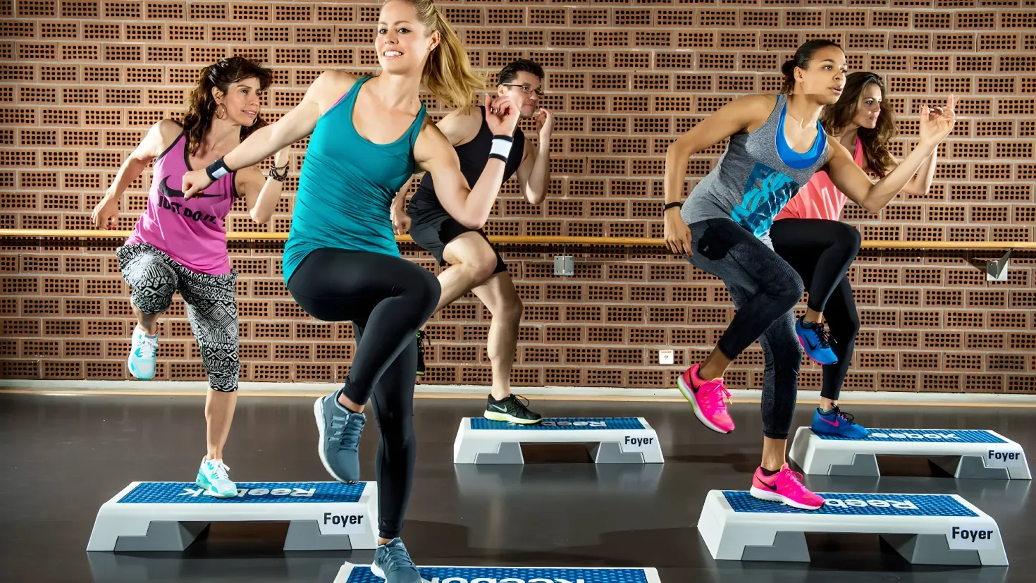 A group of women doing squats on a platform in a gym, with a wall in the background. They appear to be in the middle of a cardio workout, likely at a fitness center.