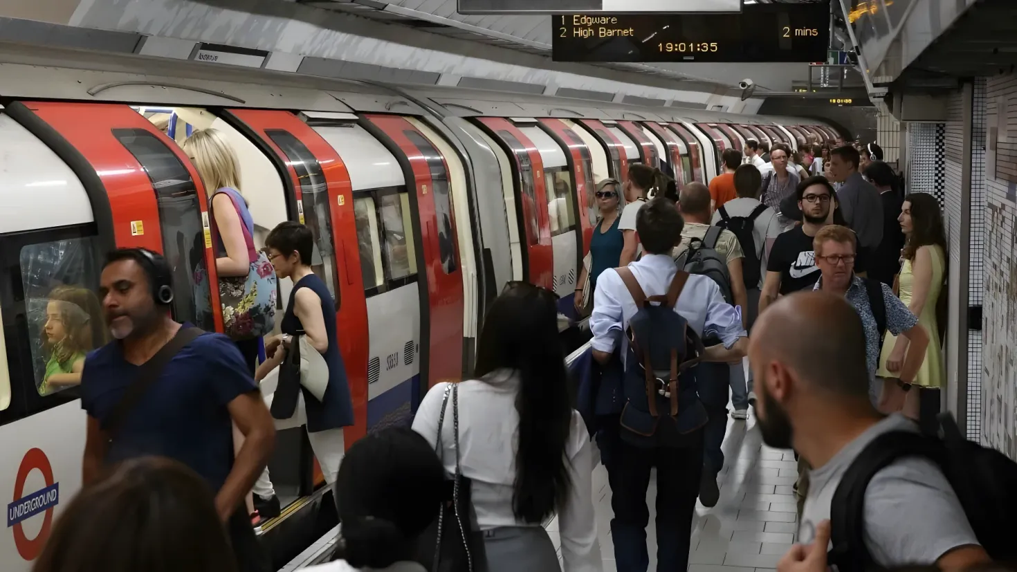 A group of people standing on a subway platform next to a train. Some of the people are wearing backpacks, suggesting they are on a journey. The train is on the left side of the image, and there are boards attached to the ceiling.