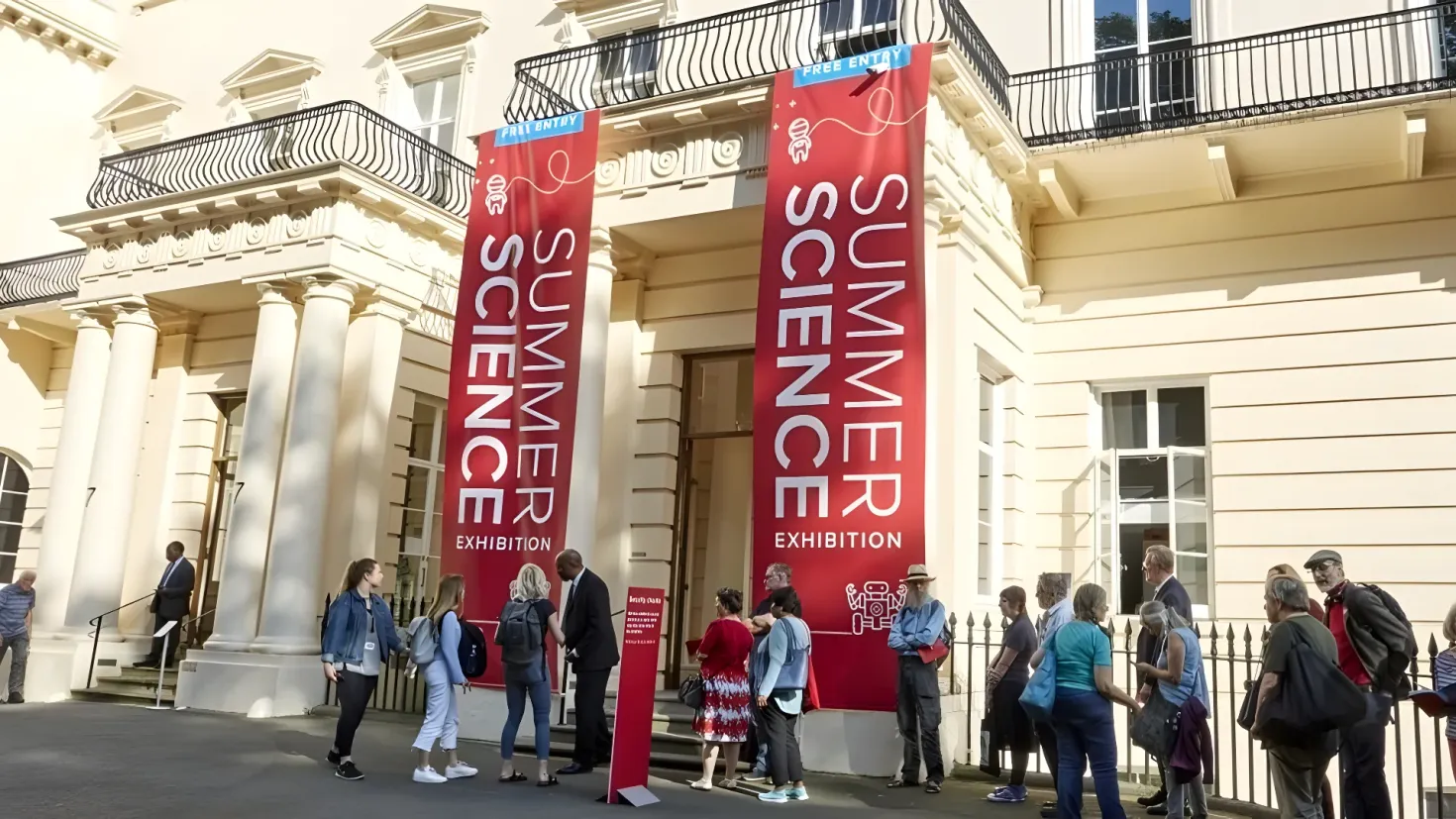 A group of people standing in front of a building with windows, railings, pillars, steps, and a banner with text on it. Some of the people are wearing hats and carrying handbags.