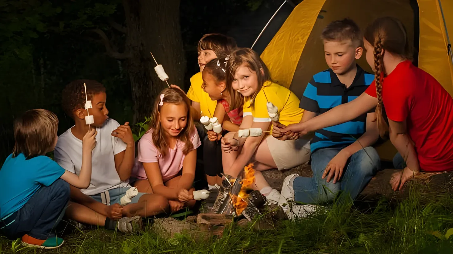 A group of children sitting around a campfire in the woods, with marshmallows in their hands. The campfire is surrounded by grass and there is a tent in the background, with trees visible in the distance.