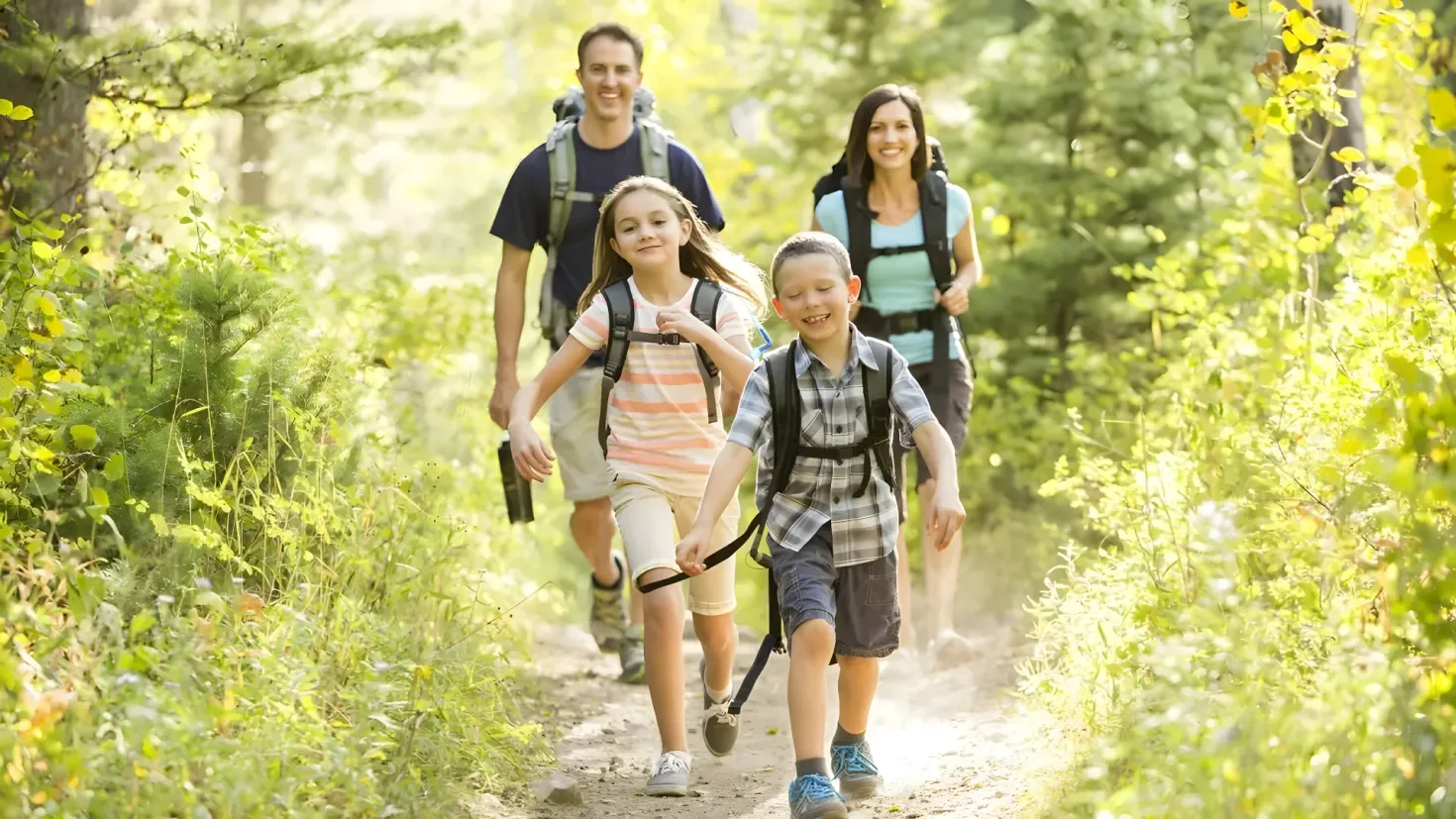 A family of four walking down a trail in the woods, wearing backpacks and smiling. The path is lined with trees on either side, creating a peaceful atmosphere.