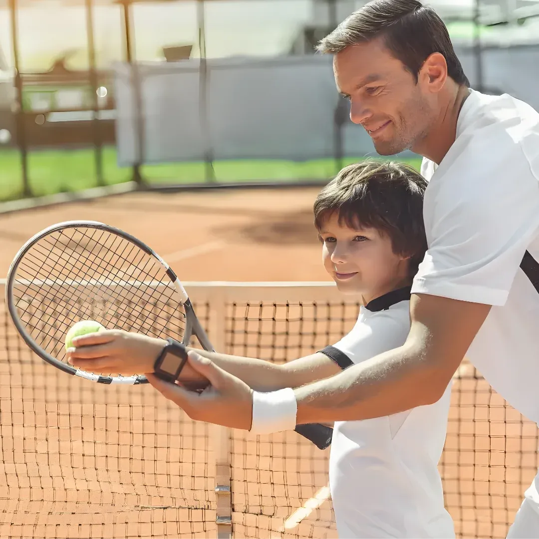 A man and a young boy playing tennis on a court. The man is holding a tennis racket and the boy is standing beside him, both of them smiling. The court is surrounded by a net, and the background is slightly blurred.