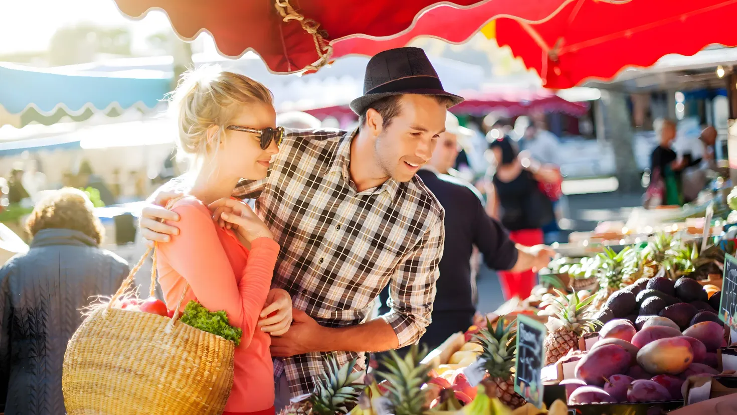 A man and woman standing in front of a fruit stand at a farmers market. The man is wearing a hat and the woman is holding a basket of vegetables. There are a number of fruits and vegetables on the table, and in the background there are a few more people and tents. The image is slightly blurred, giving it a dreamy feel.