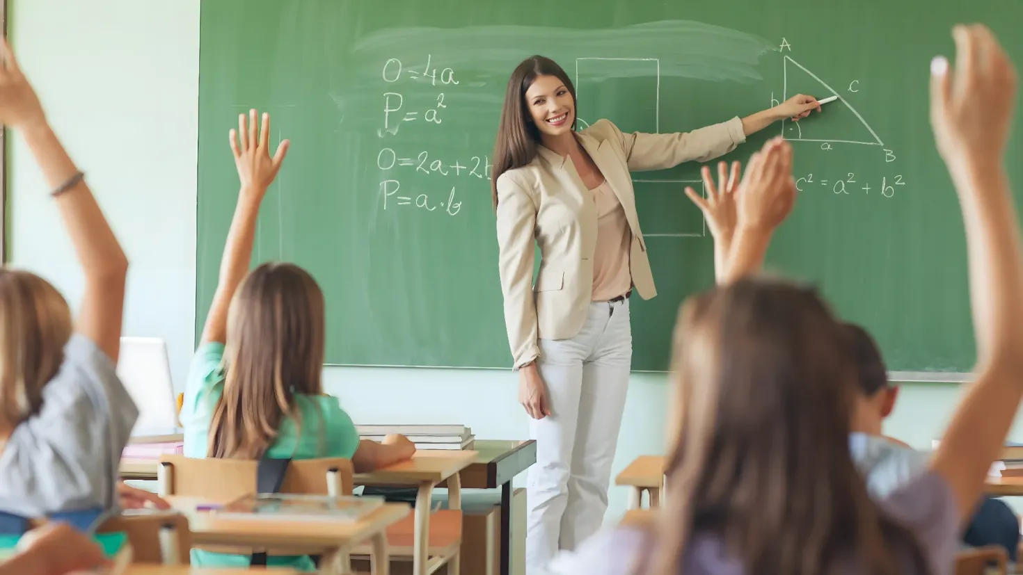 A teacher standing in front of a classroom full of students, with a smile on her face. She is holding a marker in her hand and there are books and other objects on the tables in the foreground. On the left side of the image, there is a window, suggesting that the teacher is in the middle of a lesson.