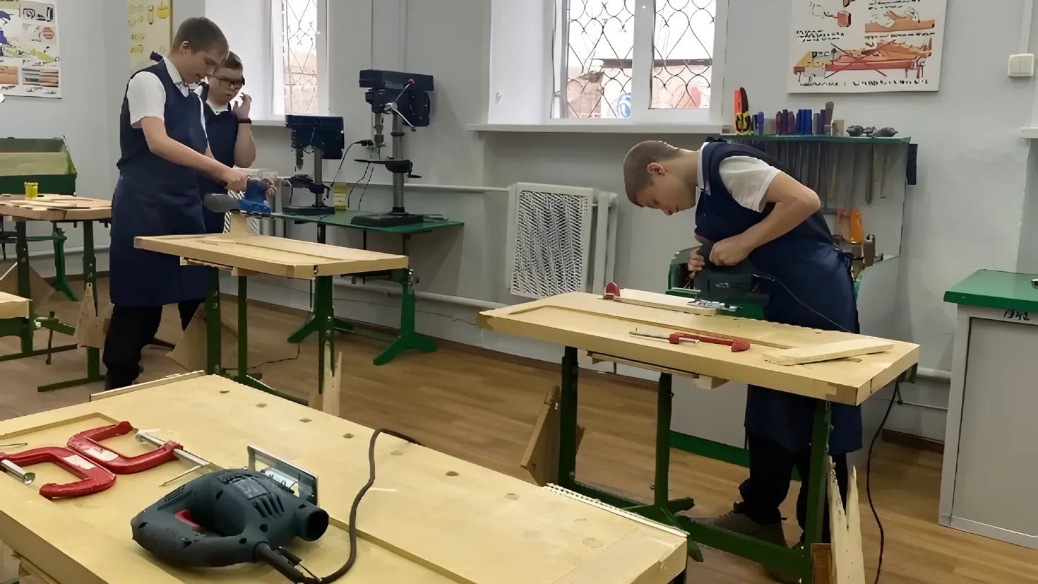 Two men working on a piece of wood in a workshop. There are tables with various tools and objects on them, as well as posters on the walls and windows in the background. On the right side of the image, there is a cupboard.