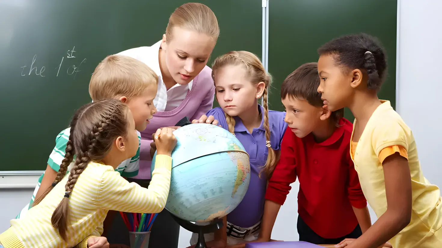 A group of children sitting around a table with a globe in front of them. On the table there are books, a pen stand with pencils, and other objects. In the background, there is a blackboard with something written on it.