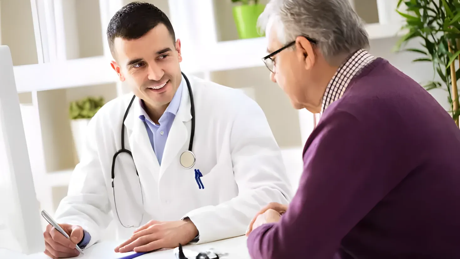 A doctor talking to an older man at a table. The doctor is wearing a stethoscope and is holding a pen, while the older man is wearing spectacles. On the table there are papers and other objects, and in the background there are plants and a wall.