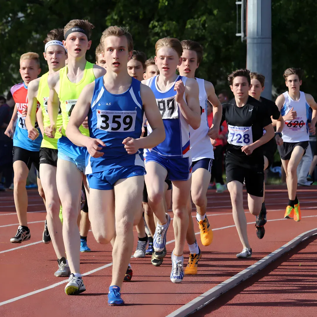 A group of young men running on a track surrounded by trees and a pole in the background. There are people sitting on the left side of the track, watching the race.