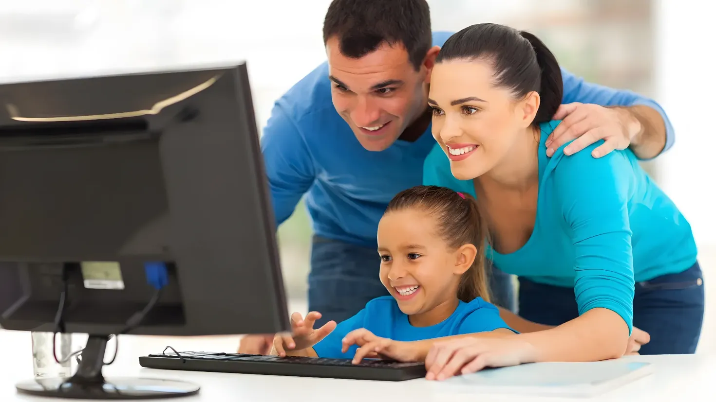 A family of three - a man, woman and a girl - looking at a computer screen with a smile on their faces. On the table in front of them is a keyboard and a monitor, suggesting they are working on a project. The background is slightly blurred, emphasizing the focus on the family and the computer.