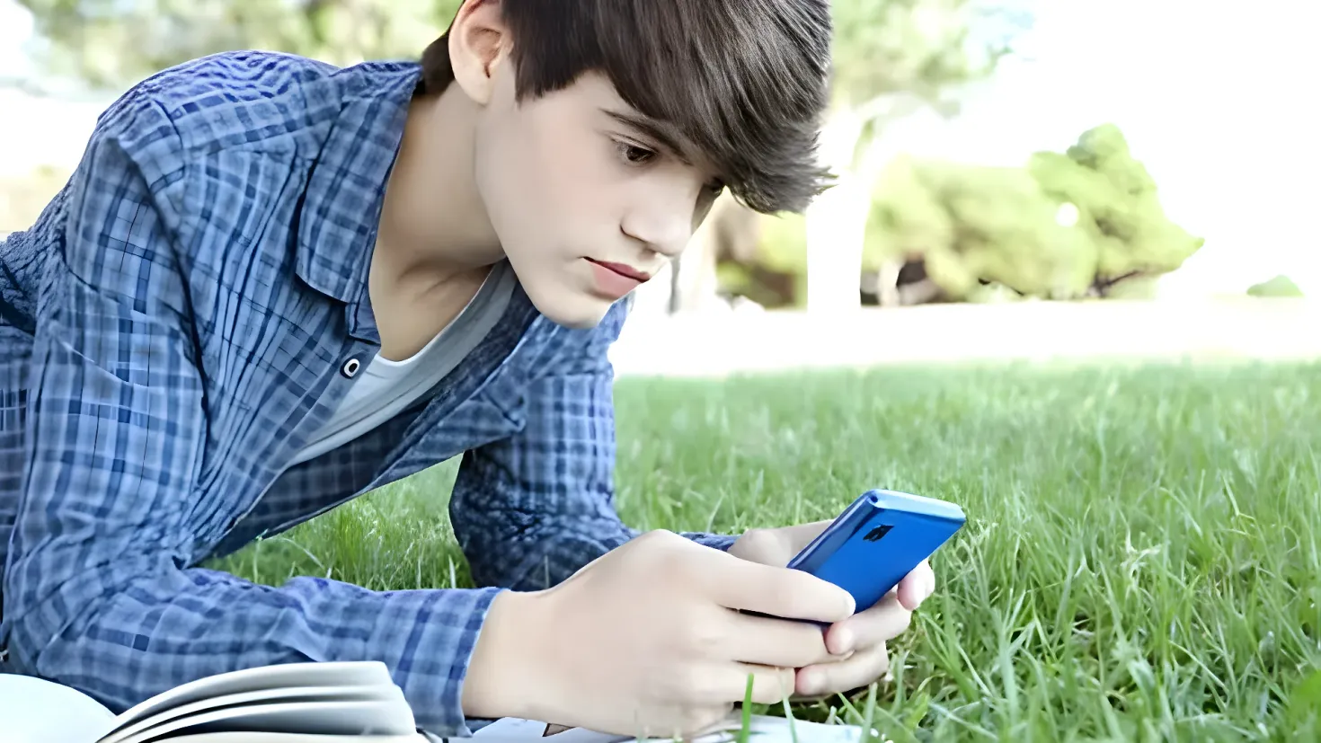 A young man laying on the grass, holding a cell phone in one hand and a book in the other. The background is slightly blurred, giving the image a dreamy feel.