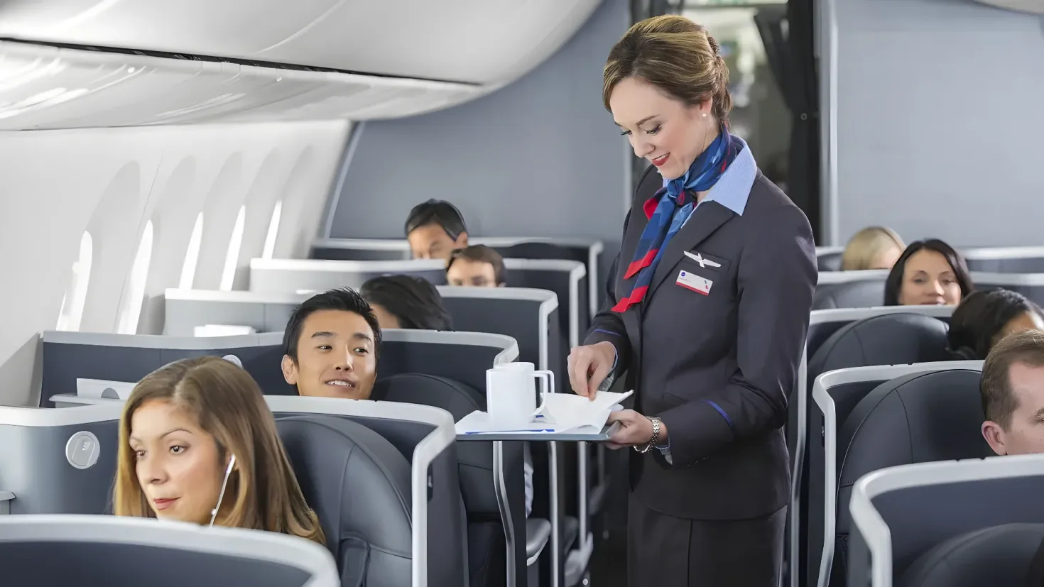 A flight attendant standing in an airplane, holding a tray with a cup and saucer on it. There are a few people sitting in the seats, and the background is slightly blurred.