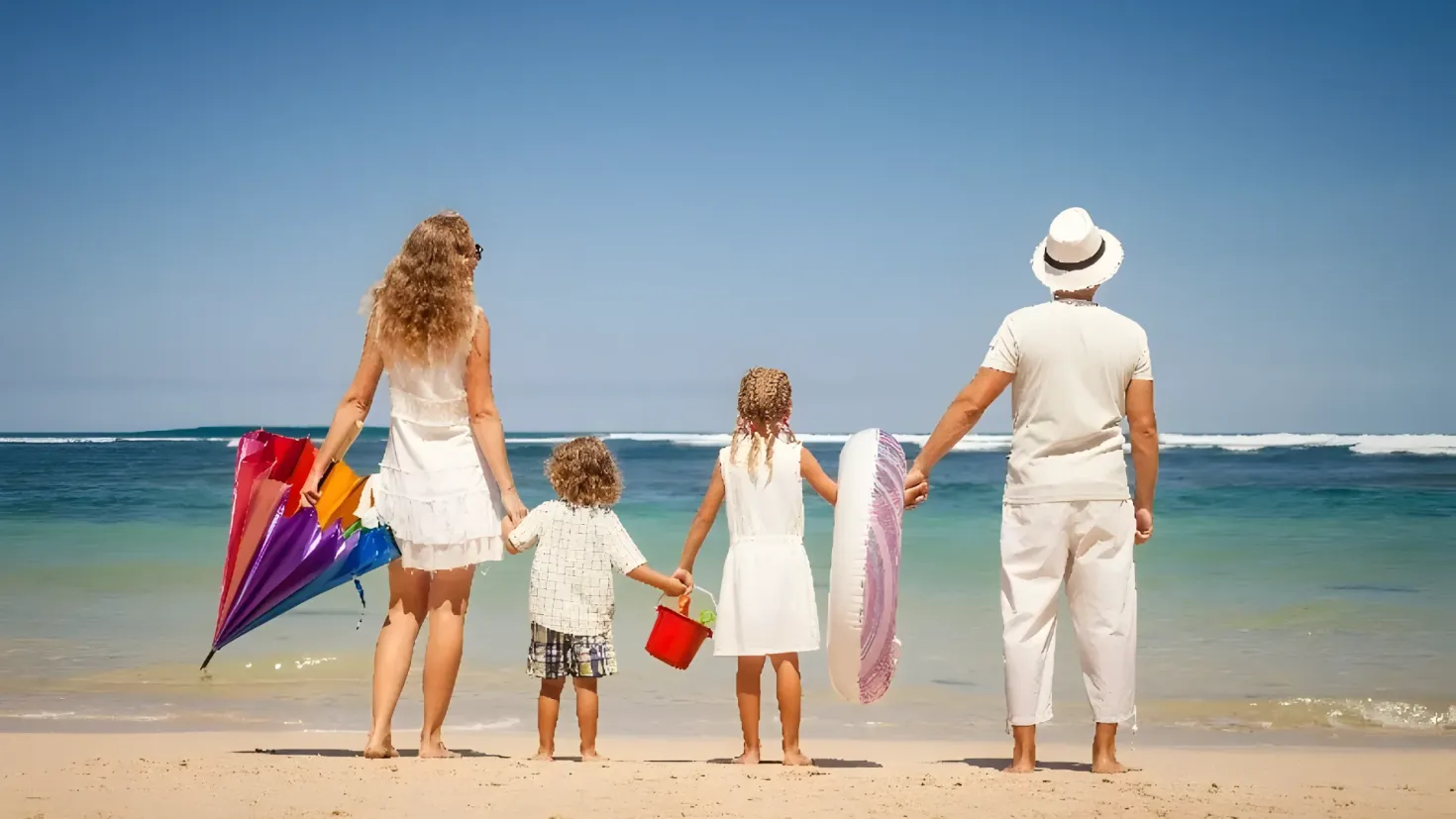 A family of four walking on the beach, with the man wearing a white dress and a hat, the woman wearing a black dress, and the two children holding colorful kites. The backdrop of the image is a large body of water and a clear blue sky.