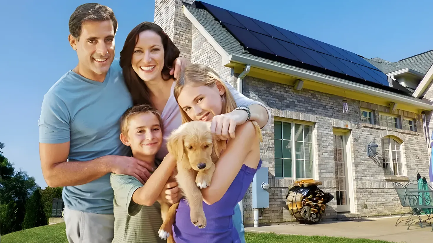 A family of four - a man, woman and two children - standing in front of a house with solar panels on the roof. The house is surrounded by lush green grass and trees, and there are chairs and a table in the foreground. The sky is visible in the background.