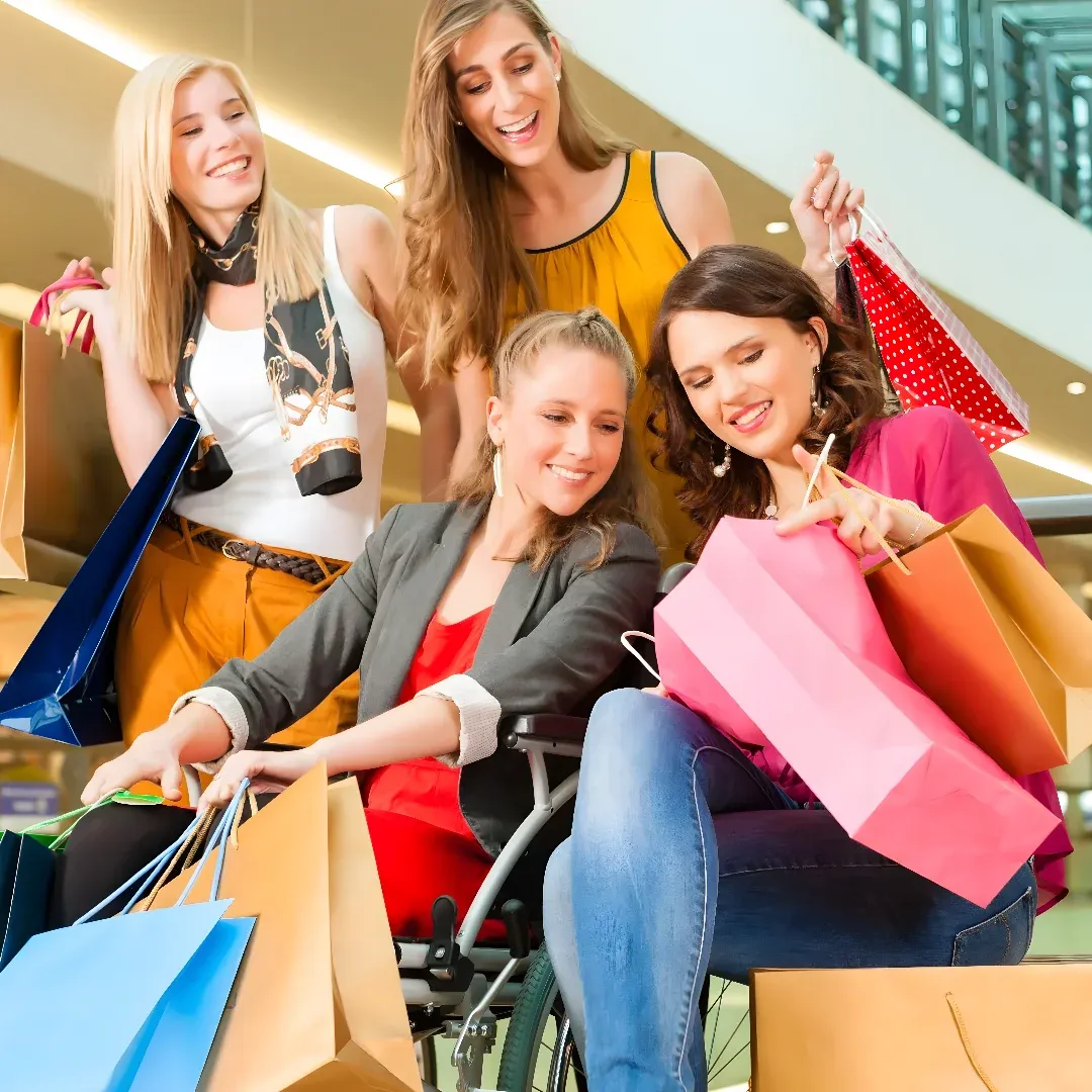 A group of four women in a shopping mall, with one woman in a wheelchair holding shopping bags. The woman in the wheelchair is wearing a red and black dress, while the other three women are wearing different colored dresses. The mall is illuminated by lights attached to the ceiling, and there is a railing in the background.