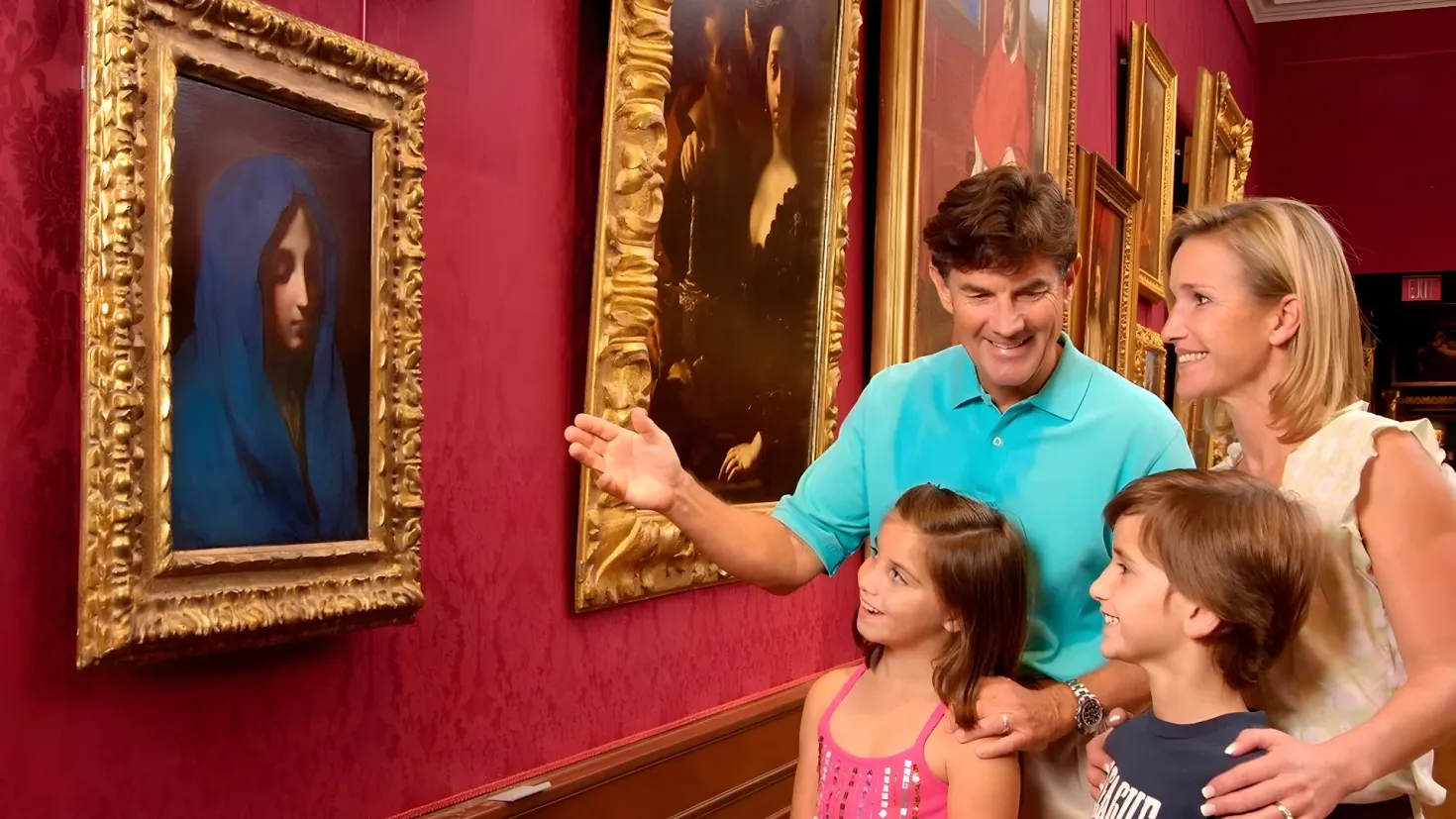 A family of four - a man, woman and two children - standing in front of a wall adorned with picture frames.