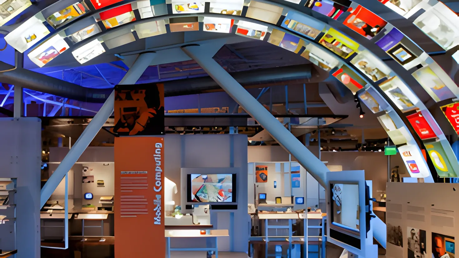 A large room filled with lots of different types of televisions, cabinetry, and a desk. There is a person standing in the room, and the walls are adorned with boards and other objects. The ceiling is illuminated with lights, creating a bright and inviting atmosphere.