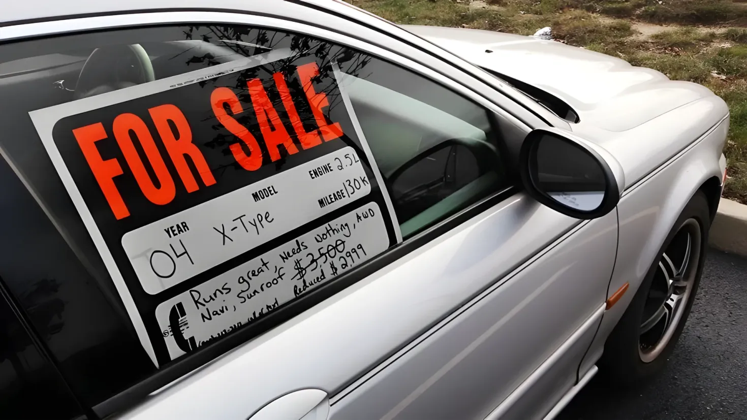 A white car parked on the side of the road with a for sale sign on the window. The car is surrounded by lush green grass and trees in the background.
