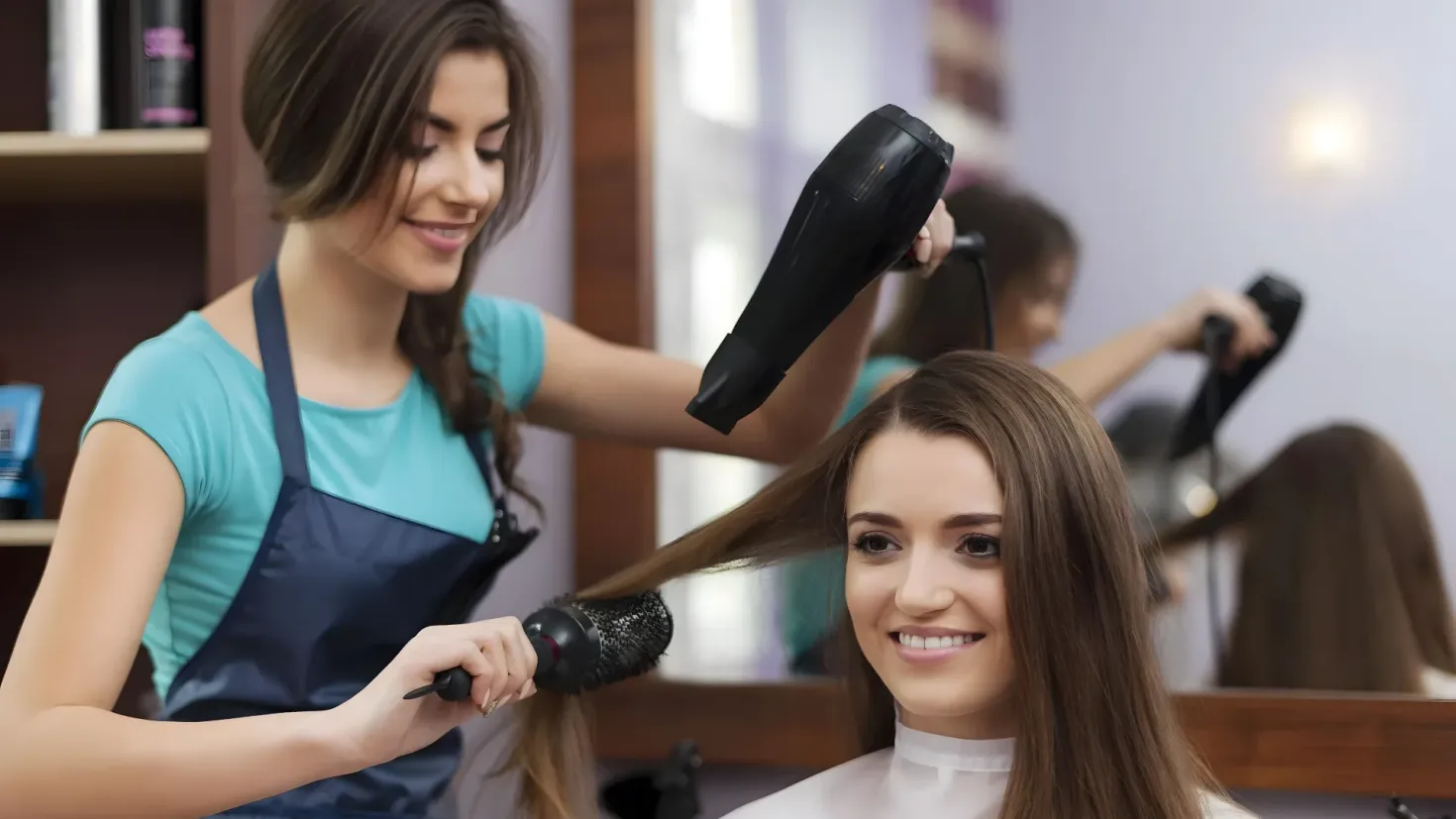 A woman getting her hair done at a salon. She is sitting in a chair with a smile on her face, while a hairdresser stands behind her, holding a hair dryer in her hand. On the left side of the image, there is a rack with various objects on it, and in the background, there are mirrors and a wall.