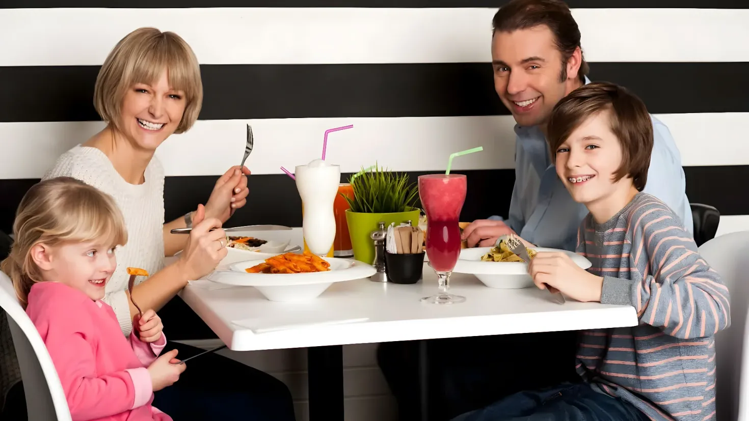 A family of four - a man, woman and two children - sitting at a table in a restaurant. On the table there are plates of food, glasses with straws, a plant in a pot, and other objects. The background of the image is a wall.