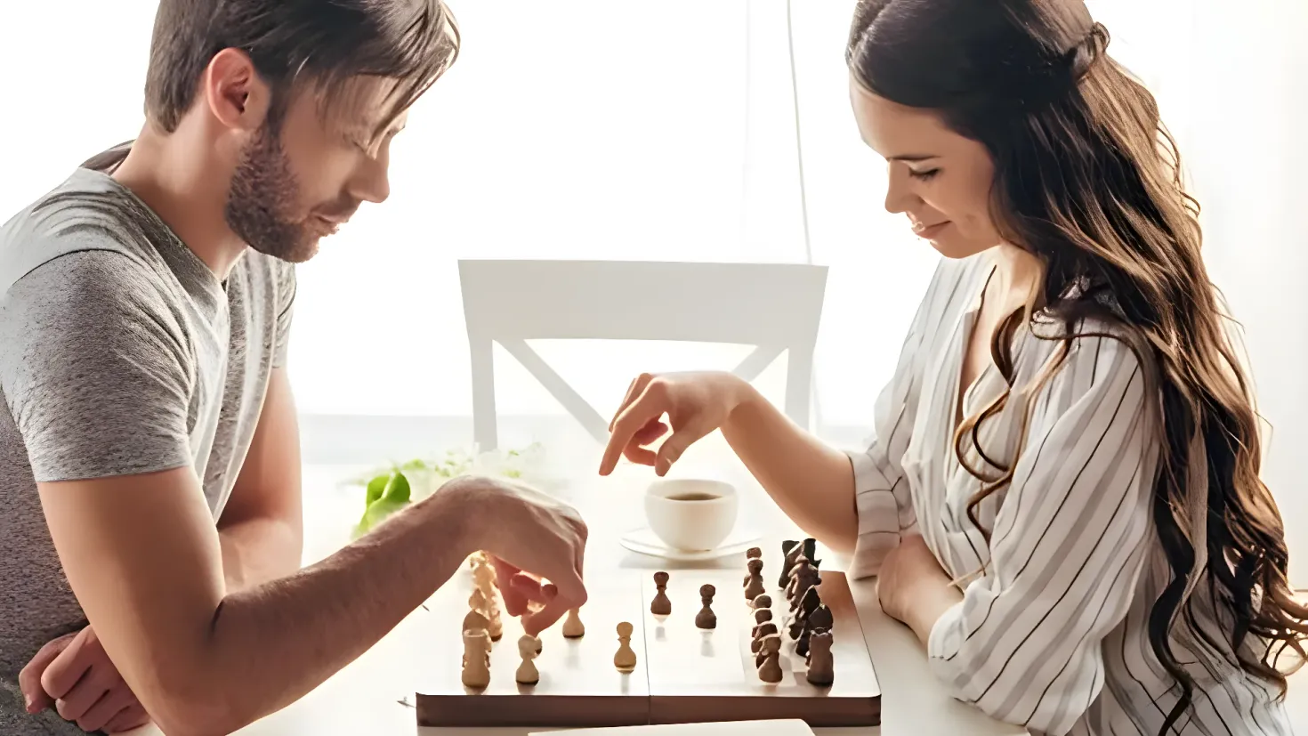 A man and woman sitting at a table playing chess. On the table there are chess pieces, a cup with saucer, and other objects. In the background there is a chair and a window.