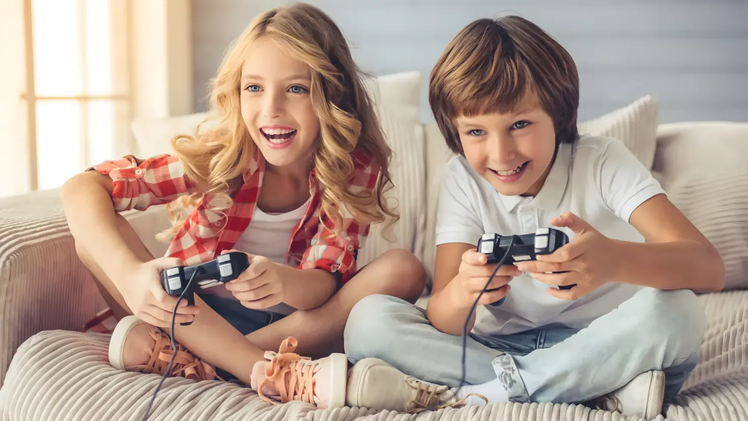 Two children sitting on a couch, each holding a video game controller in their hands. They are surrounded by cushions and there is a wall and a window in the background.