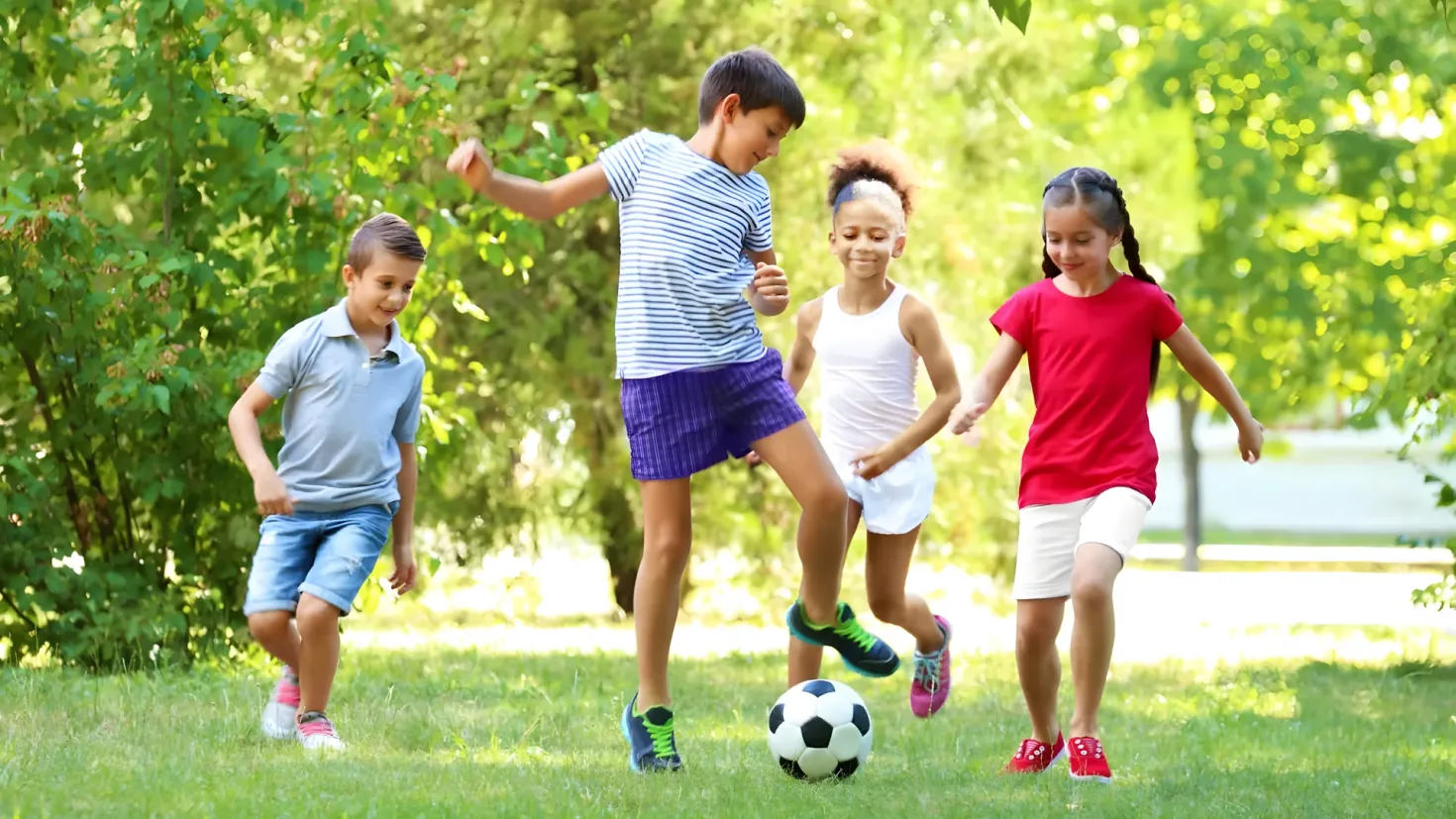 A group of children playing soccer in a park, surrounded by lush green grass and trees in the background.