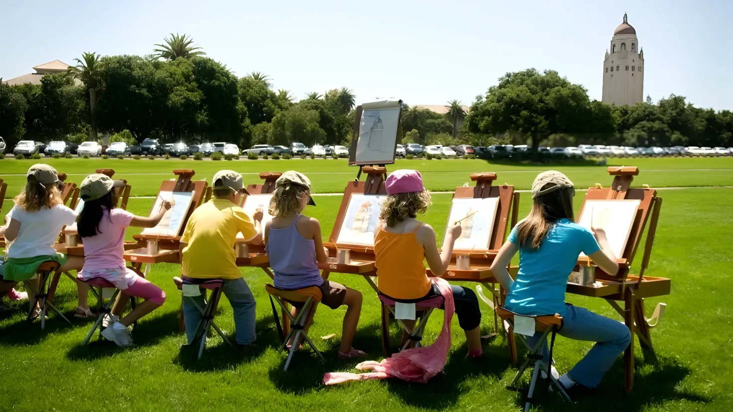 A group of people sitting at easels in front of a building, surrounded by trees, vehicles, and a clear blue sky. On the easels, there are boards with paintings, and the ground is covered in lush green grass.