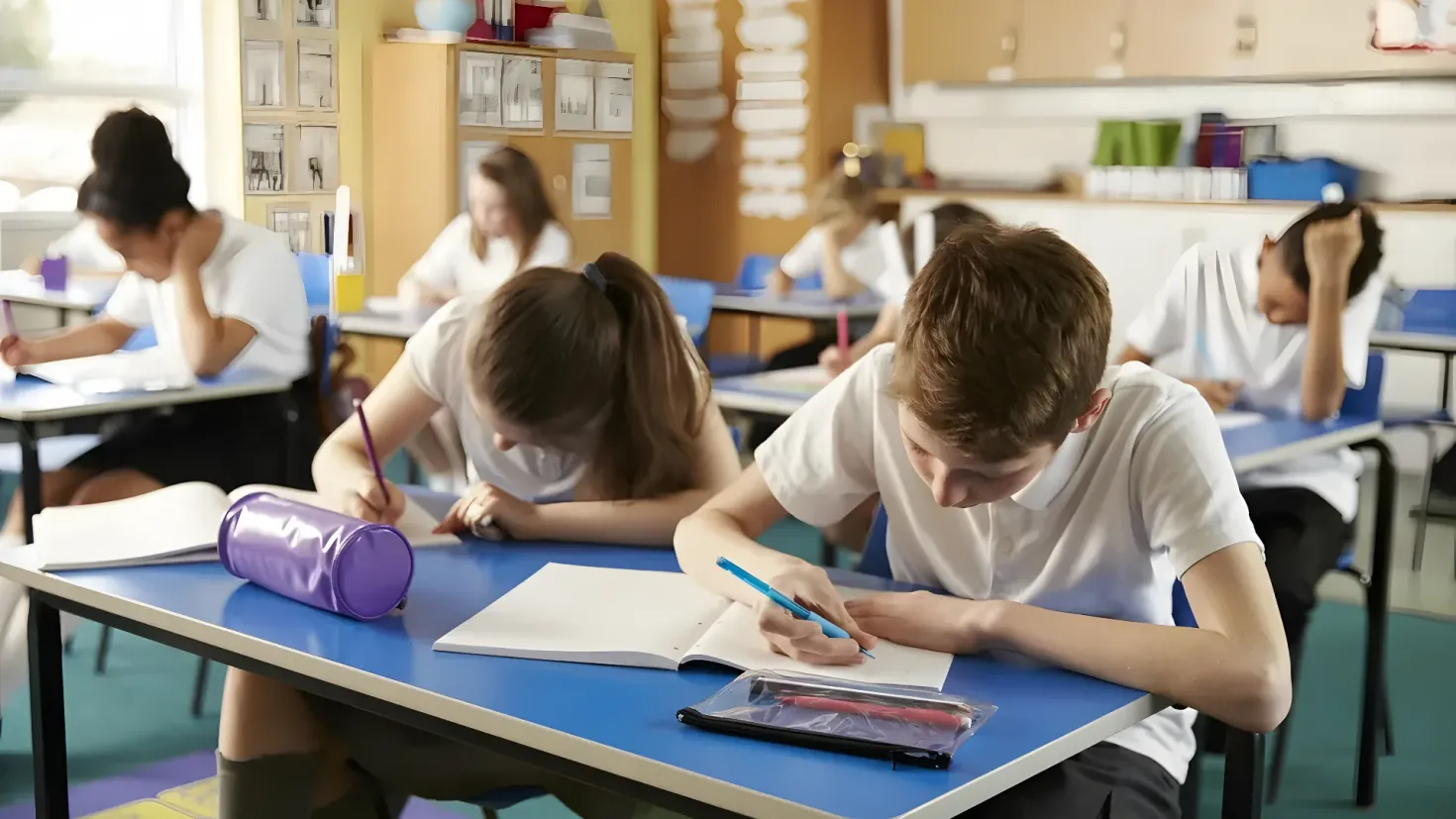 A group of students sitting at desks in a classroom, with books, pens, and other objects on the tables. In the background, there are cupboards and a wall, and on the left side of the image there is a window.