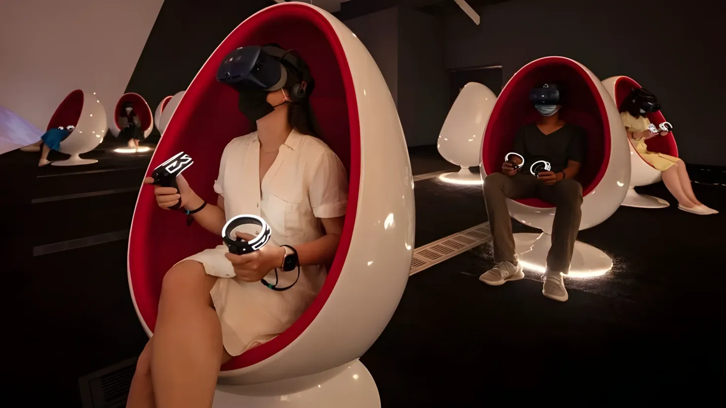 A woman sitting in a red egg-shaped chair with a remote control in her hand. She is wearing a white dress and a black helmet, and is surrounded by other people sitting in the same chair. The background of the image is a white wall.
