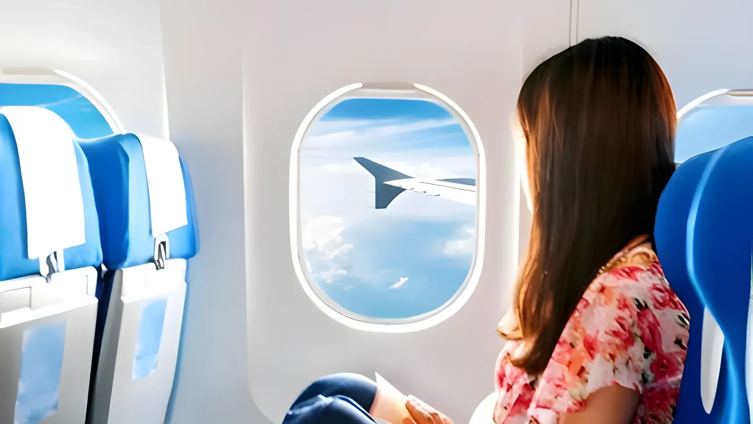 A woman sitting in an airplane, looking out the window at the sky. She is holding a book in her hands, and the windows of the plane are visible in the background.