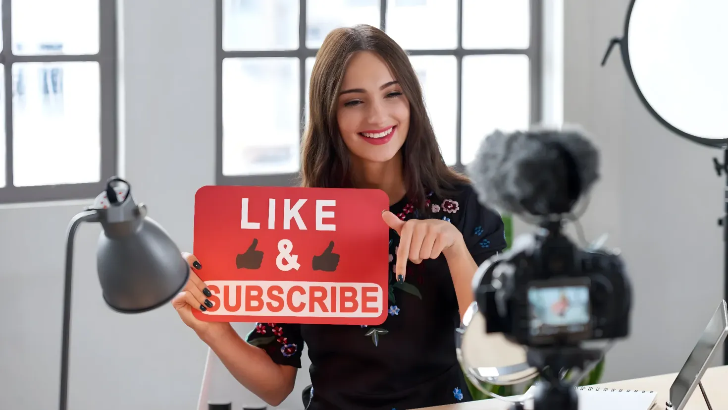 A woman holding up a sign that says "Like & Subscribe" in front of a camera, a laptop, a book, a lamp, and other objects on a table. In the background, there are windows and a wall, suggesting that the woman is in an office setting. This image is likely related to social media marketing, as it is a reminder of the importance of taking the necessary steps to make the most of your business.