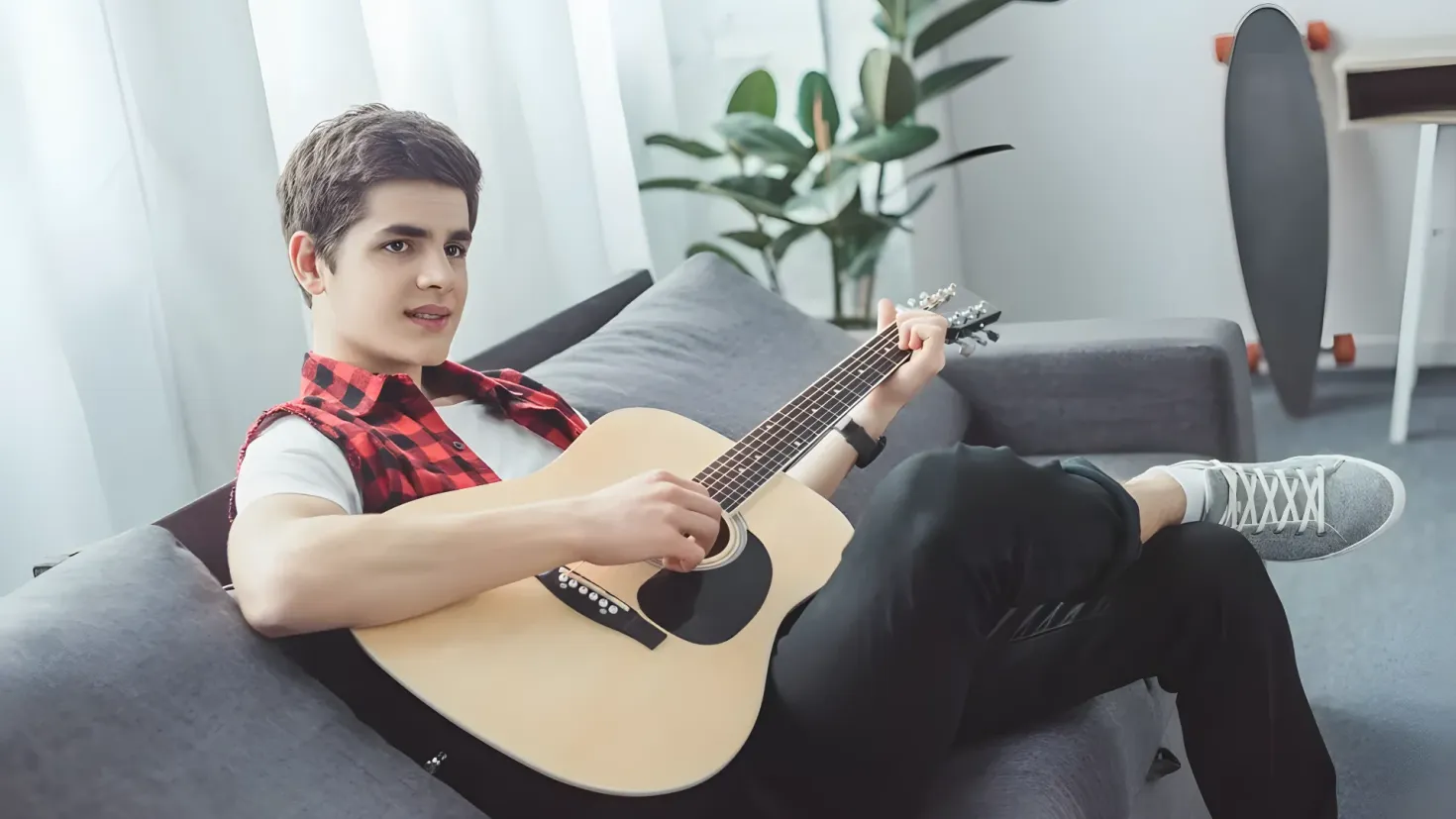 A young man sitting on a couch, playing an acoustic guitar. He is surrounded by a houseplant, a stool, and curtains, creating a cozy atmosphere.