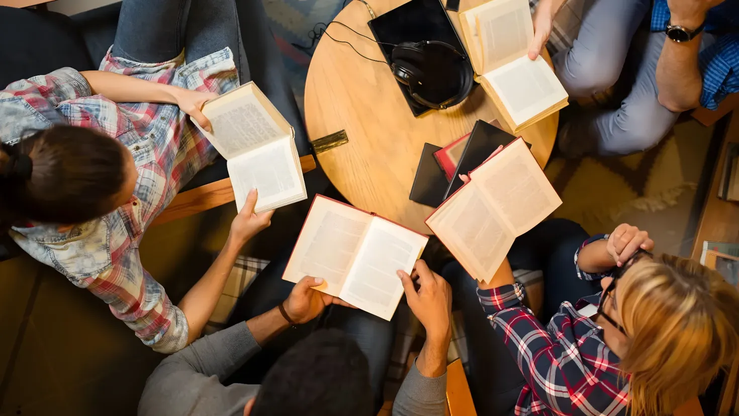 A group of people sitting around a table, each holding a book in their hands. On the table there are books, a headset, and other objects. To the right of the table is a cupboard filled with books.