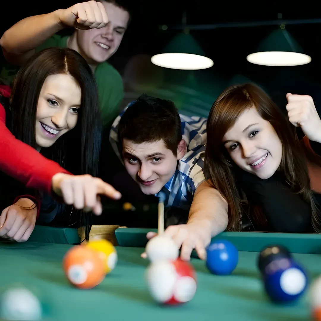 A group of four young people playing snooker in a pool hall, illuminated by lights from the ceiling. They are all smiling and appear to be having a great time.
