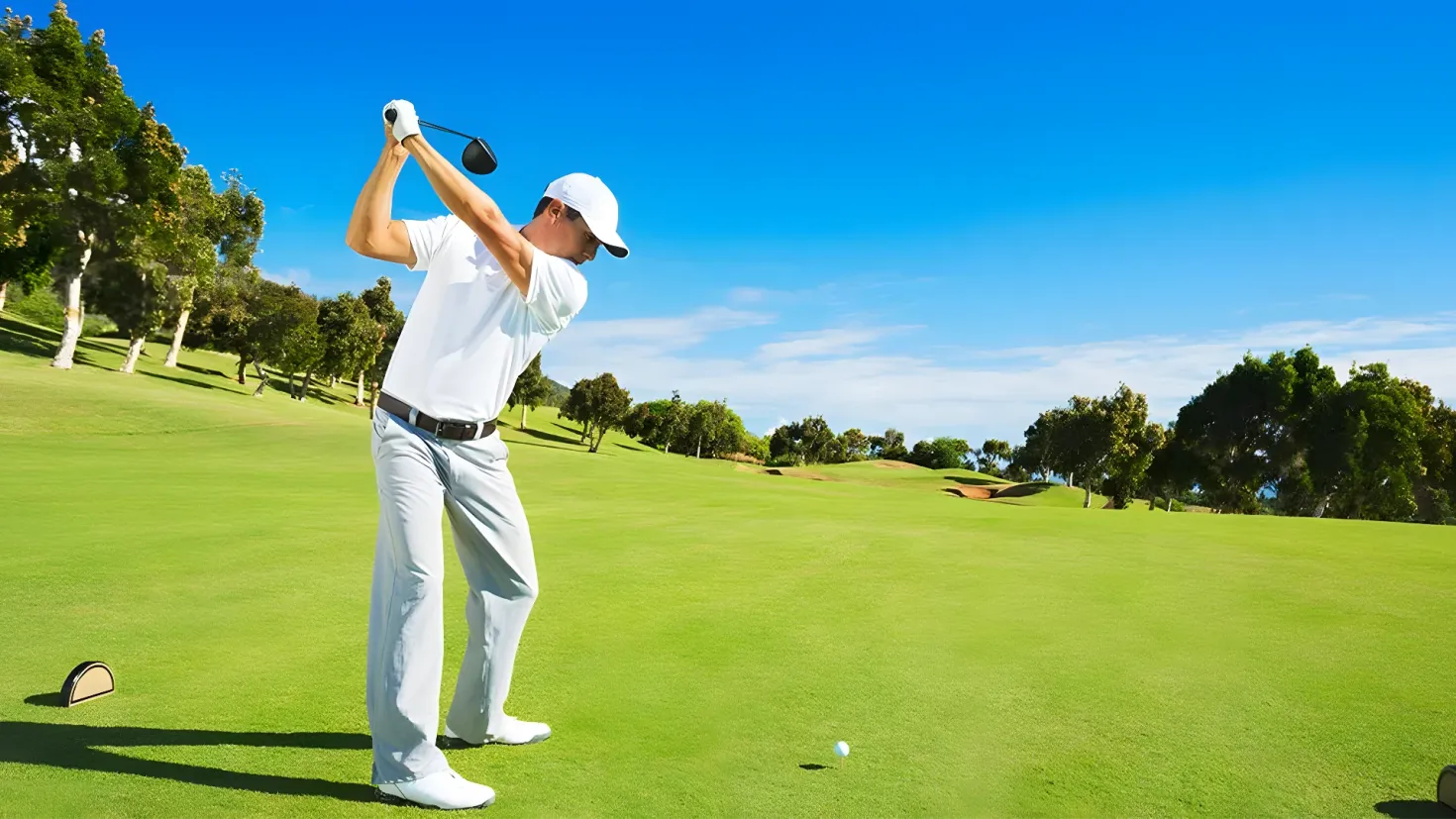 A man wearing a white t-shirt, white pants, and a white cap, swinging a golf club on top of a lush green field. In the background, there are trees and a clear blue sky.