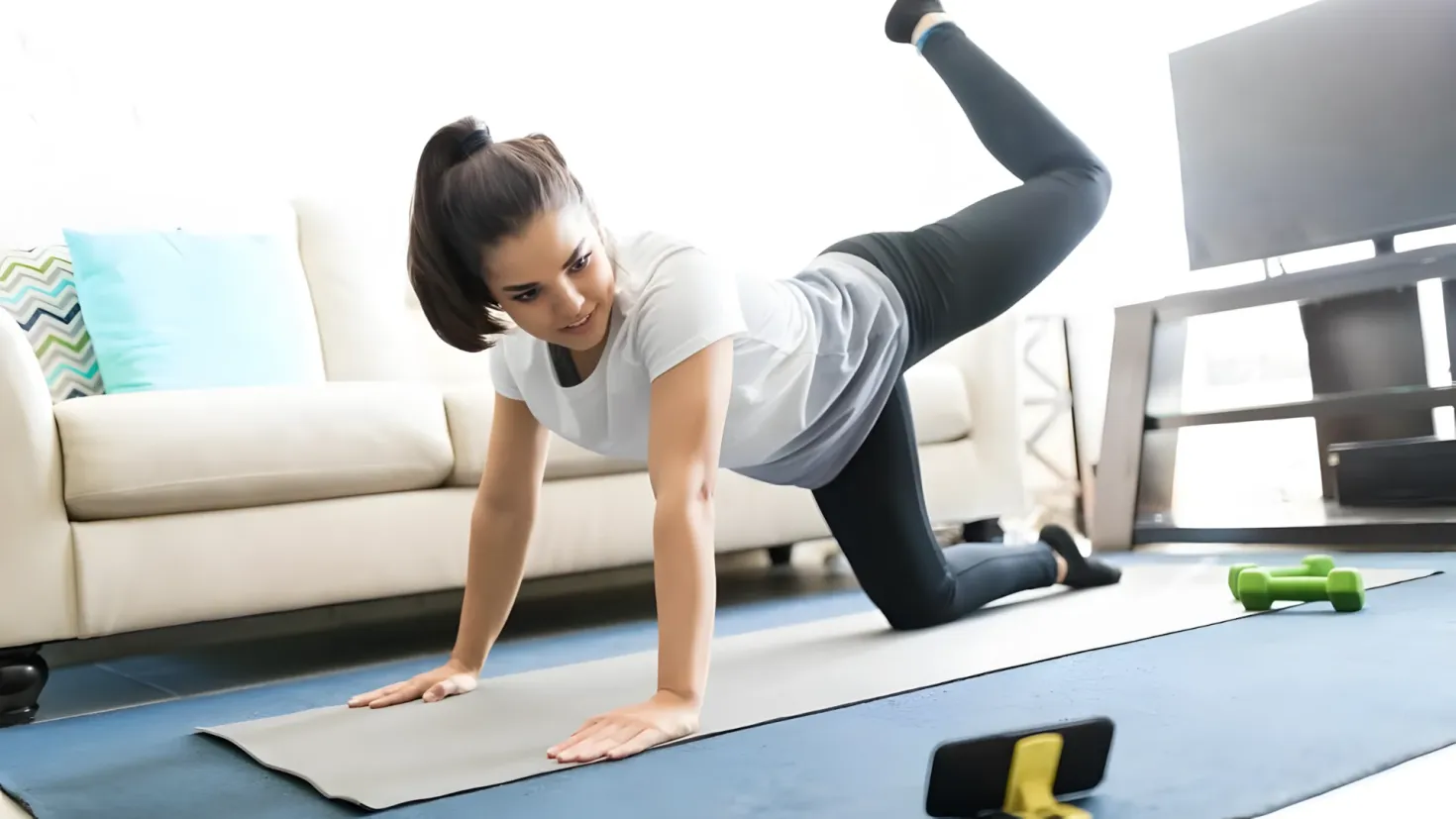 A woman doing push ups on a yoga mat in front of a television, with dumbbells on the floor and a sofa with pillows in the background.
