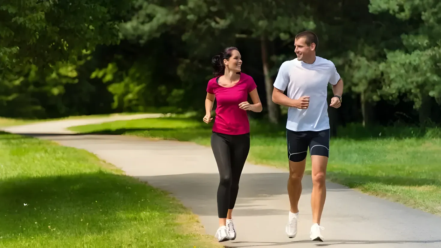 A man and woman jogging down a path in a park, surrounded by lush green grass and tall trees. They are both wearing sneakers, suggesting they are taking part in a healthy lifestyle.